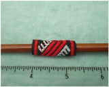 Dreadz Red Black and White patterned Dreadlock Fabric Hair Cuff, code #3144, measuring 1 and 1 quarter inches long by half an inch wide, shown on brown wooden knitting needle behind transparent rule demonstrating length