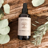 A 200 millilitre bottle of RAW ROOTs ACV Apple Cider Vinegar Herbal Dreadlock Hair Rinse shown laid upon the bark of a tree surrounded by some leaves