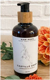 A 250 millilitre bottle of Mandarin and Vanilla scented RAW ROOTS Natural Organic Castille Shampoo/Soap displayed on a wooden table top with flowers in front and behind